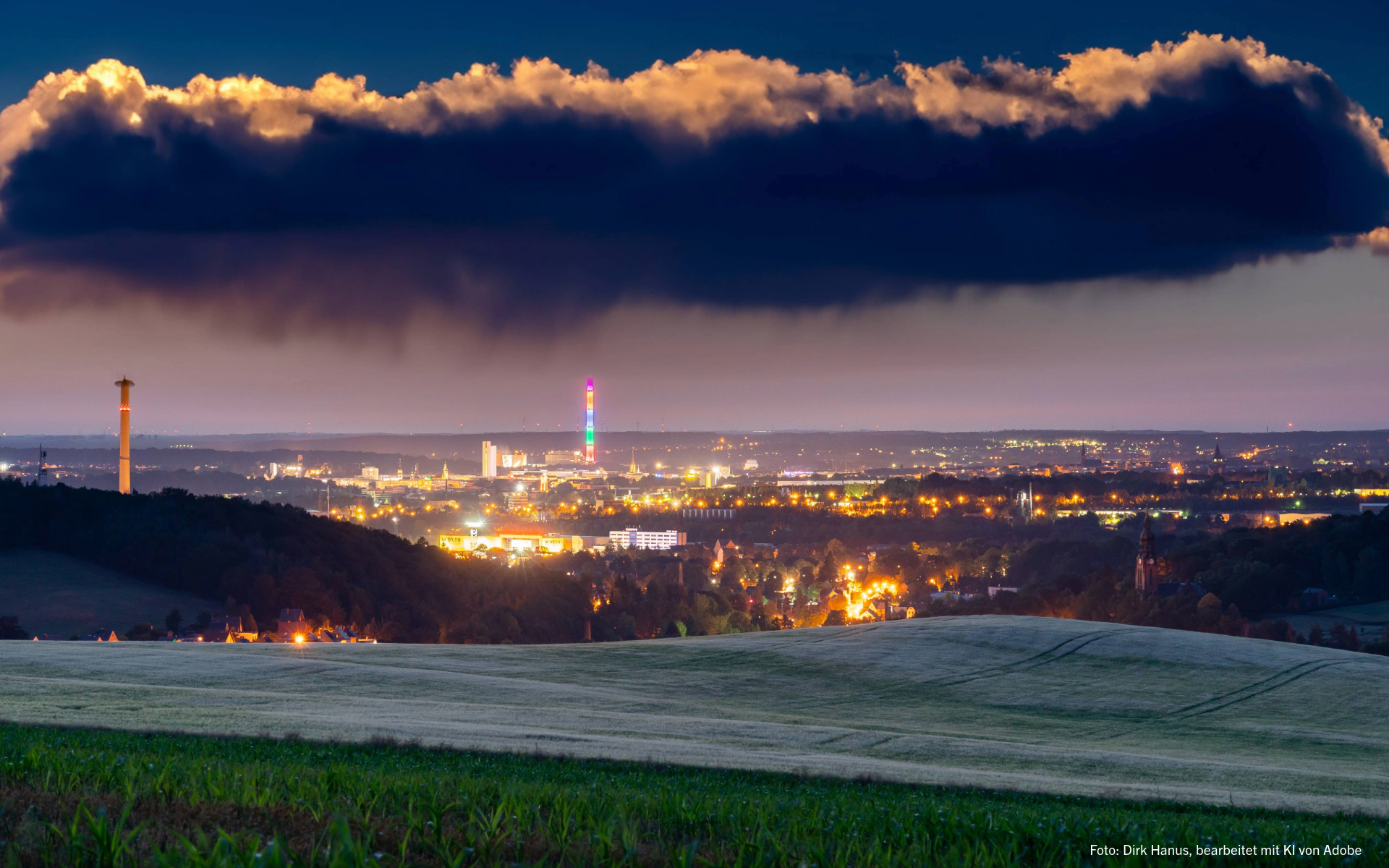 Das Bild zeigt den Blick über das abendliche und beleuchtete Chemnitz mit dunklen Regenwolken. 
